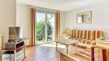A spacious living room with a sofa, a TV on a stand, a table, and a sliding glass door leading to a balcony.