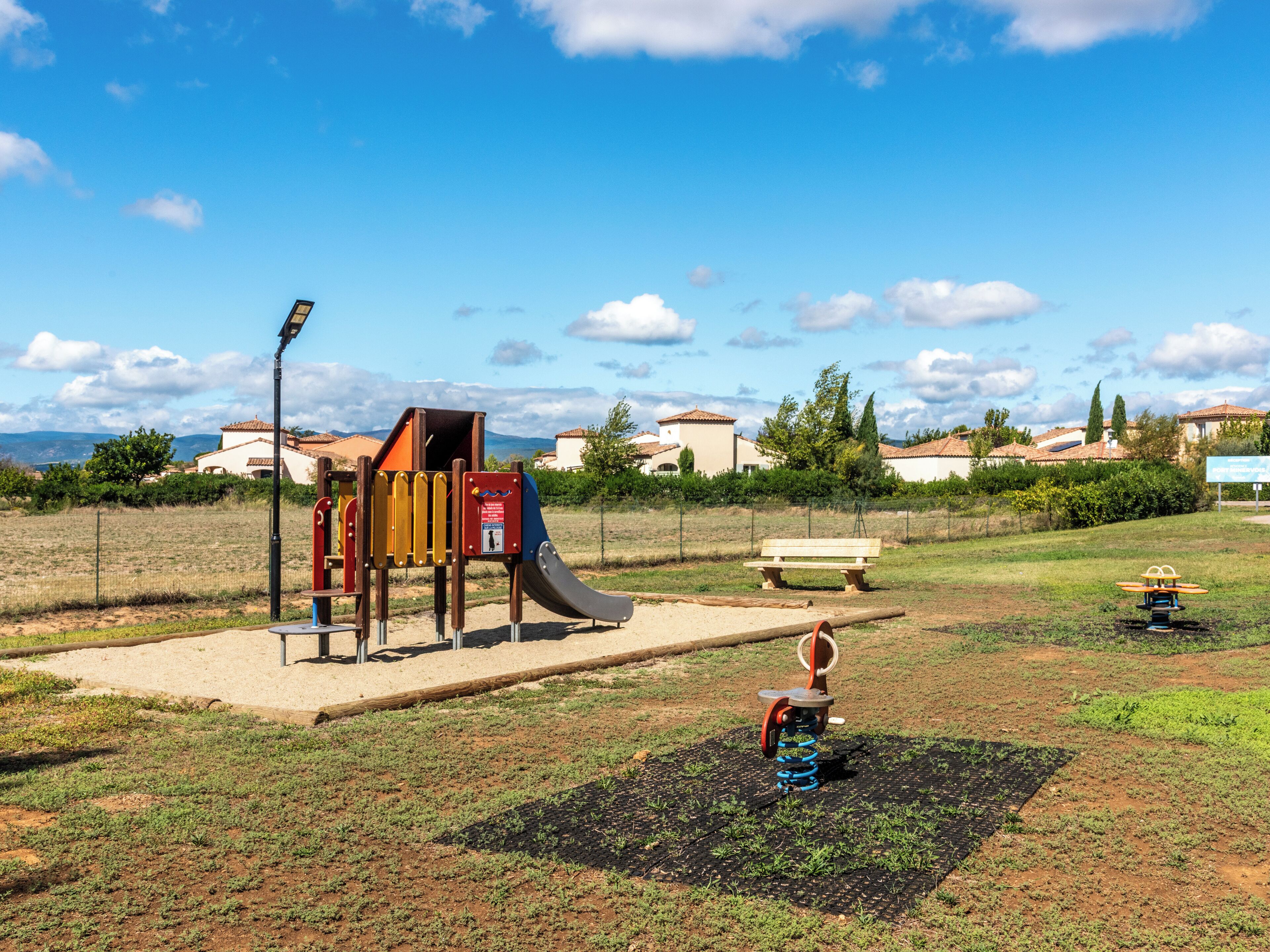 Une aire de jeux avec un toboggan et une structure d’escalade, entourée d’une zone herbeuse délimitée par une clôture, avec des bancs et une vue sur les maisons au loin.