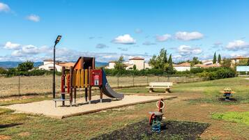 Une aire de jeux avec un toboggan et une structure d’escalade, entourée d’une zone herbeuse délimitée par une clôture, avec des bancs et une vue sur les maisons au loin.