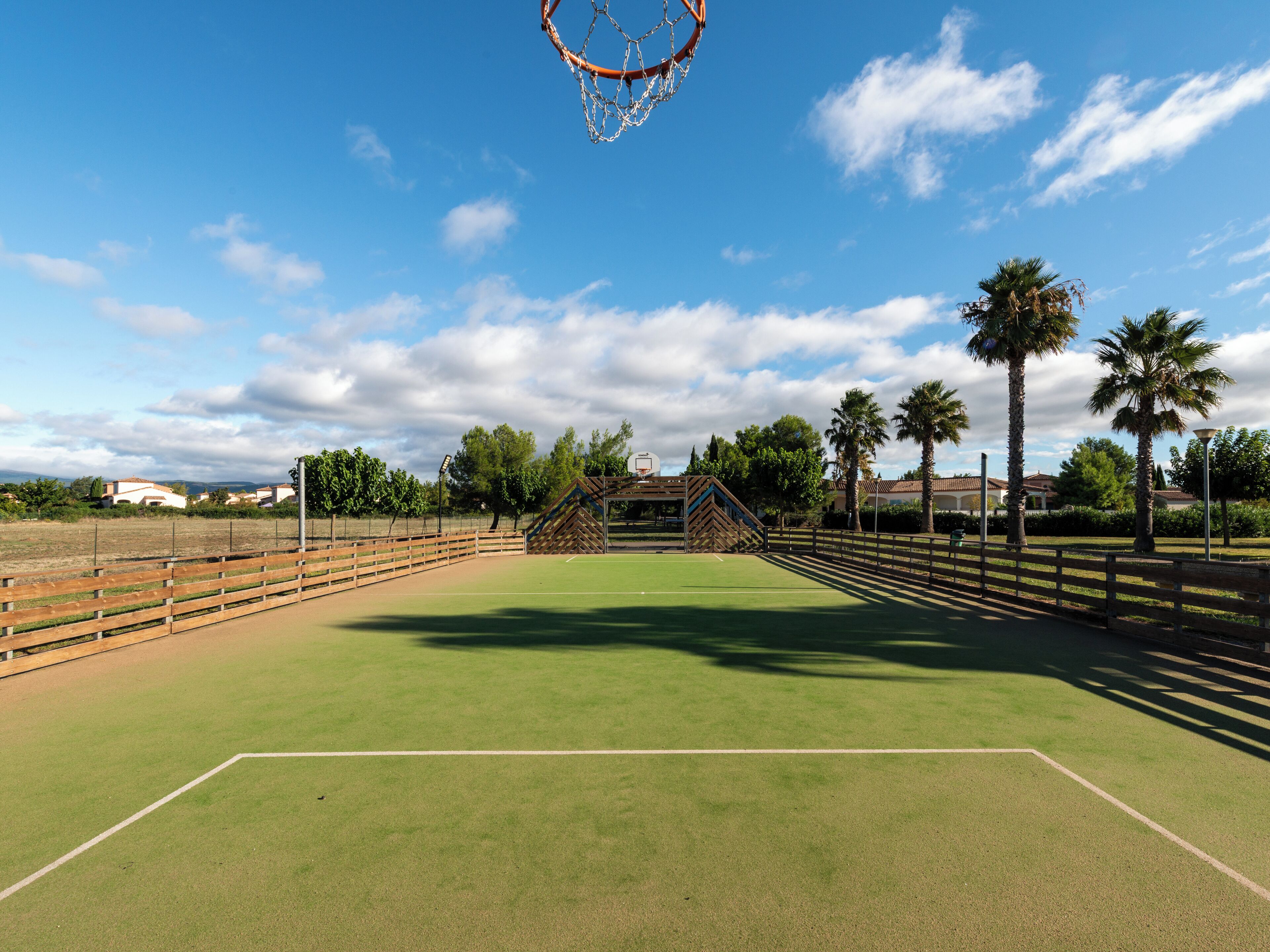 Een buitenbasketbalveld met een houten omheining, een basketbalring en palmbomen op de achtergrond.