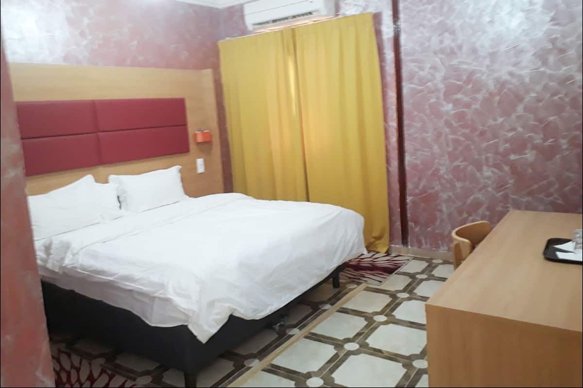 A hotel room with a bed, a desk, a chair, a window with curtains, and an air conditioning unit.