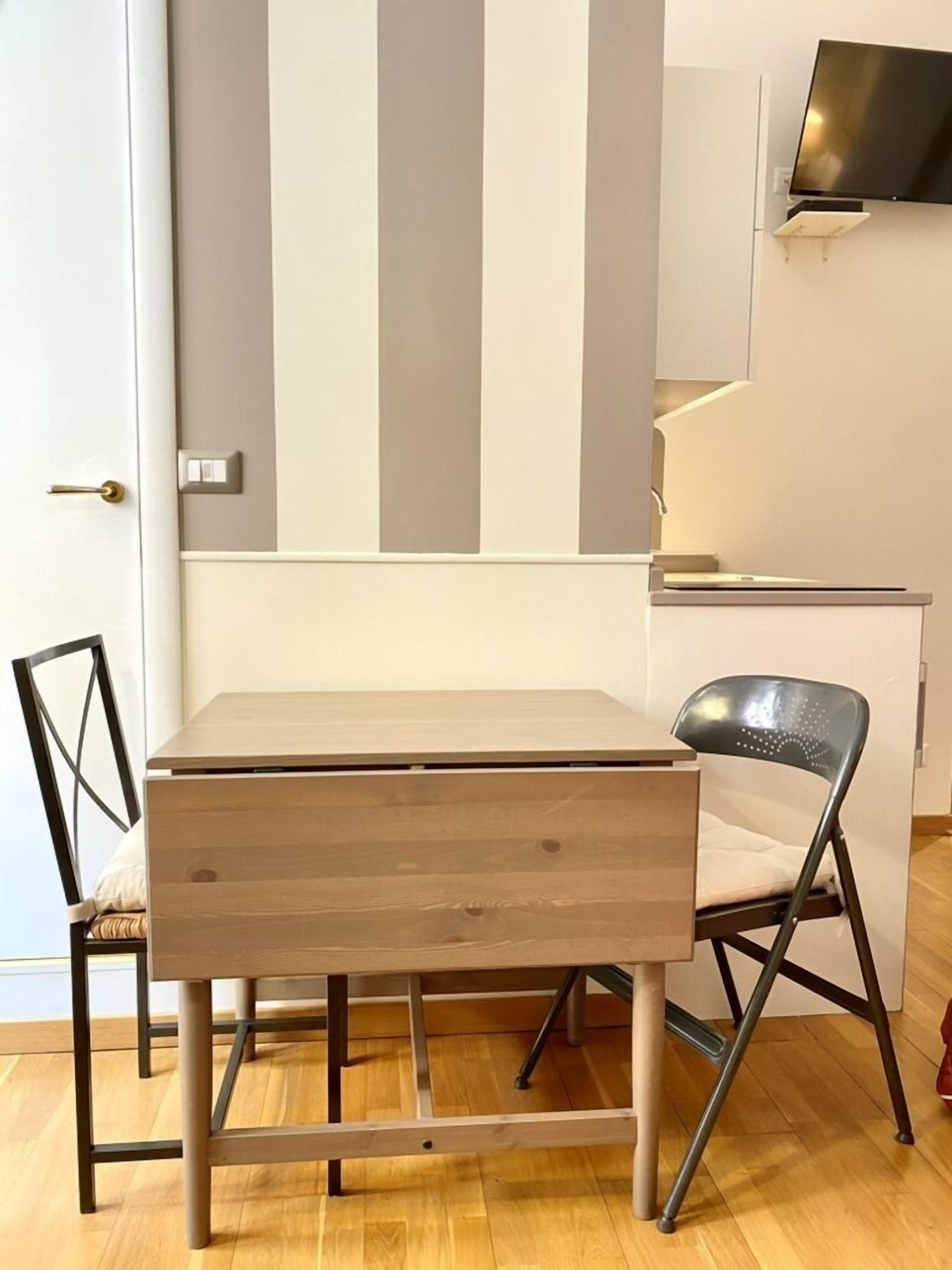 Une chambre moderne avec une table et des chaises en bois, un téléviseur à écran plat fixé au mur et un escalier menant à un niveau supérieur.