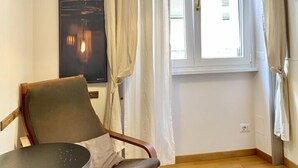 A room with a wooden floor, a chair, a small round table, a hanging lamp, a window with curtains, and a painting on the wall. - Roome B&B (Rome)
