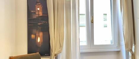Ein Zimmer mit Holzboden, einem einzelnen Sessel, einem kleinen runden Tisch, einer Stehlampe und einem fenster mit Vorhängen.