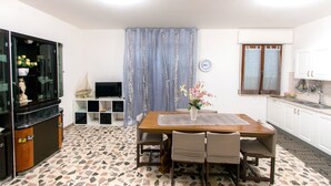 A dining area with a wooden table and chairs, a television on a stand, a clock on the wall, and a kitchen area with cabinets and a sink. - B&B Casa Anna Maria (Porto Sant'Elpidio)