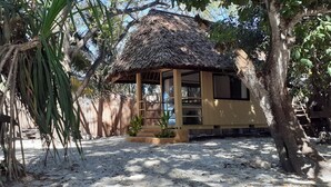 A thatched-roof hut with a sandy floor, surrounded by trees and plants. - Mikadi Beach Camp (Dar es Salaam)