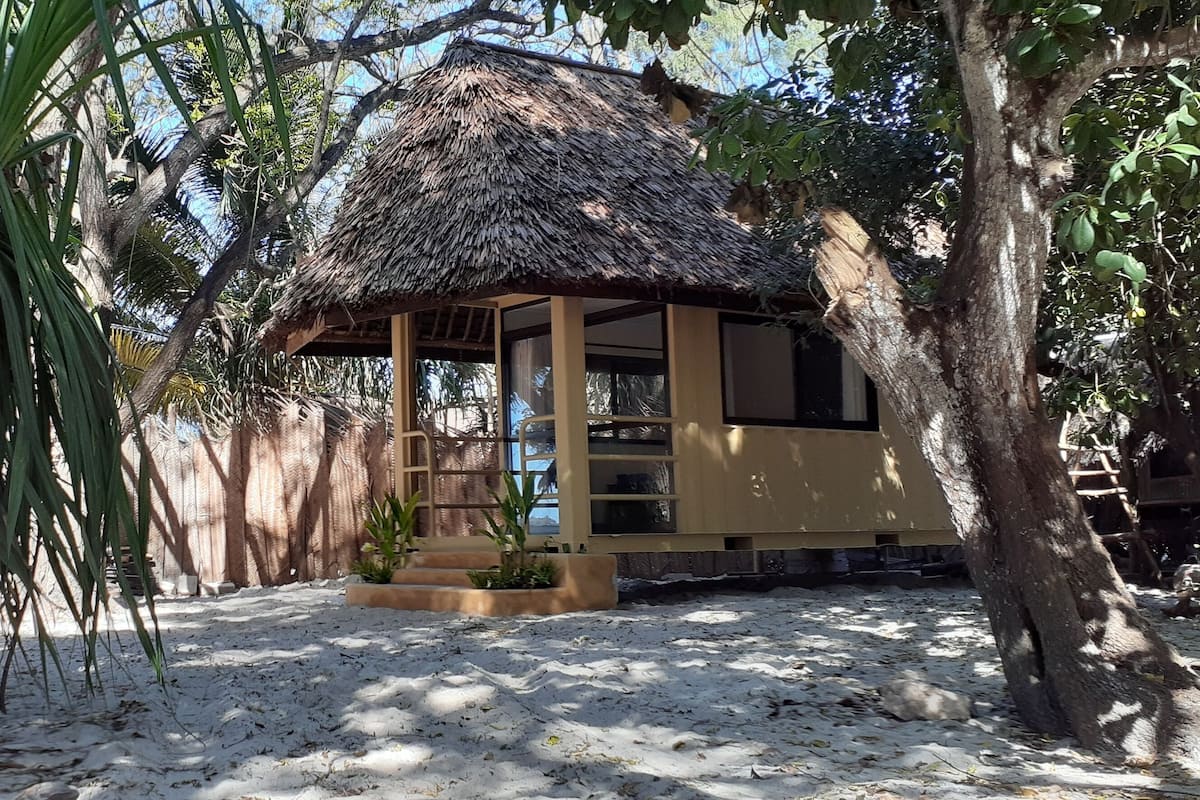 Une hutte au toit de chaume, avec un sol sablonneux, entourée d’arbres et de plantes.