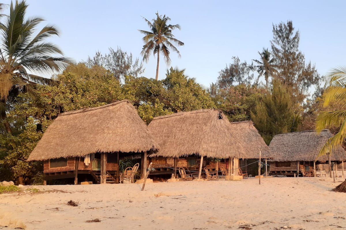 Des huttes à toits de chaume, construites en bois, entourées de palmiers et d’autres plantes, sur une plage de sable.