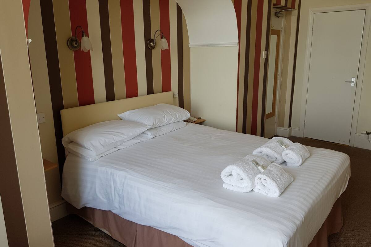 Een netjes opgemaakt bed met witte lakens en twee opgerolde handdoeken, een telefoon op het nachtkastje, en een muur met verticale strepen.