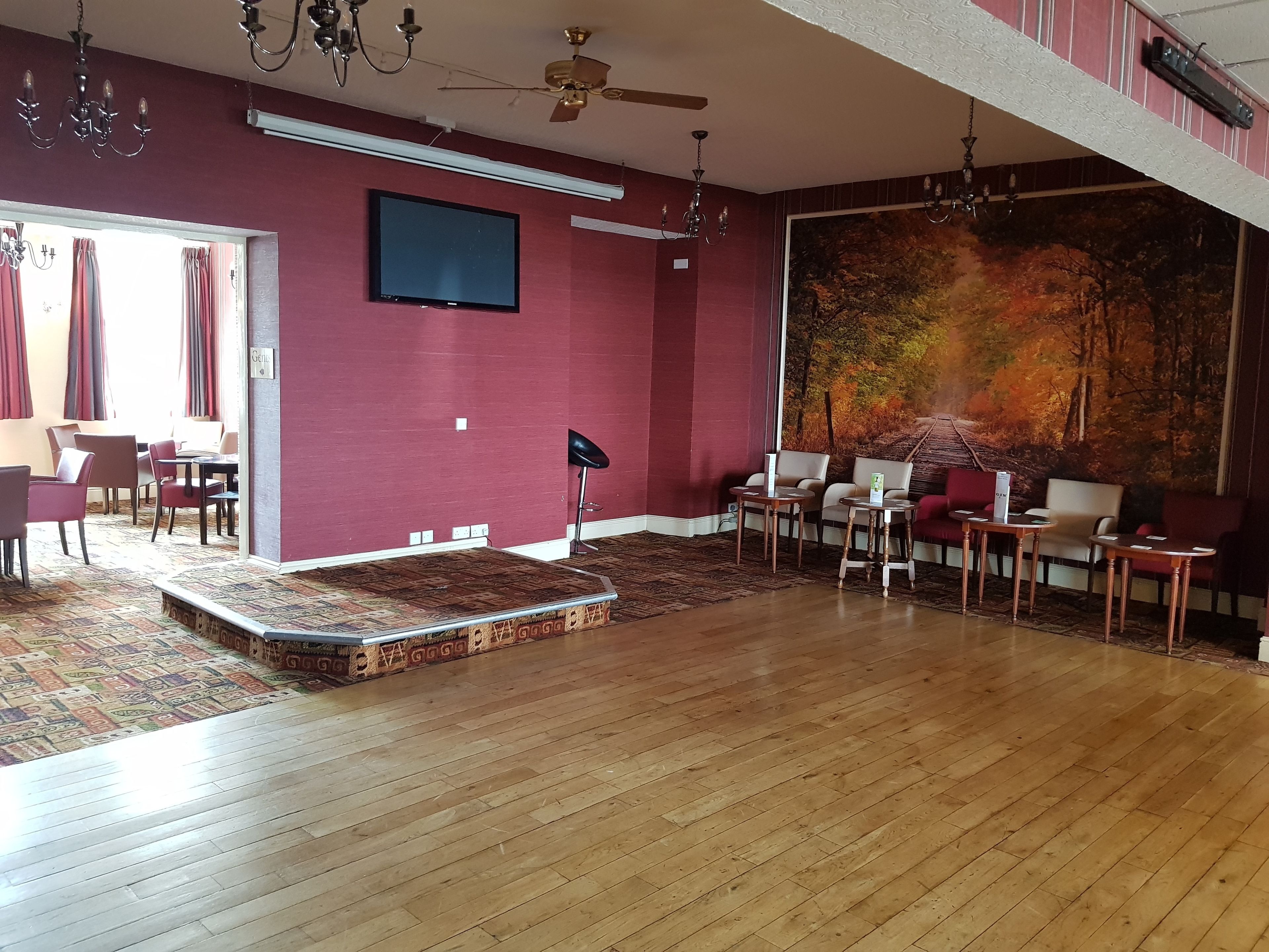 Un amplio salón con piso de madera, paredes rojas, un televisor montado y un gran mural con motivos otoñales.