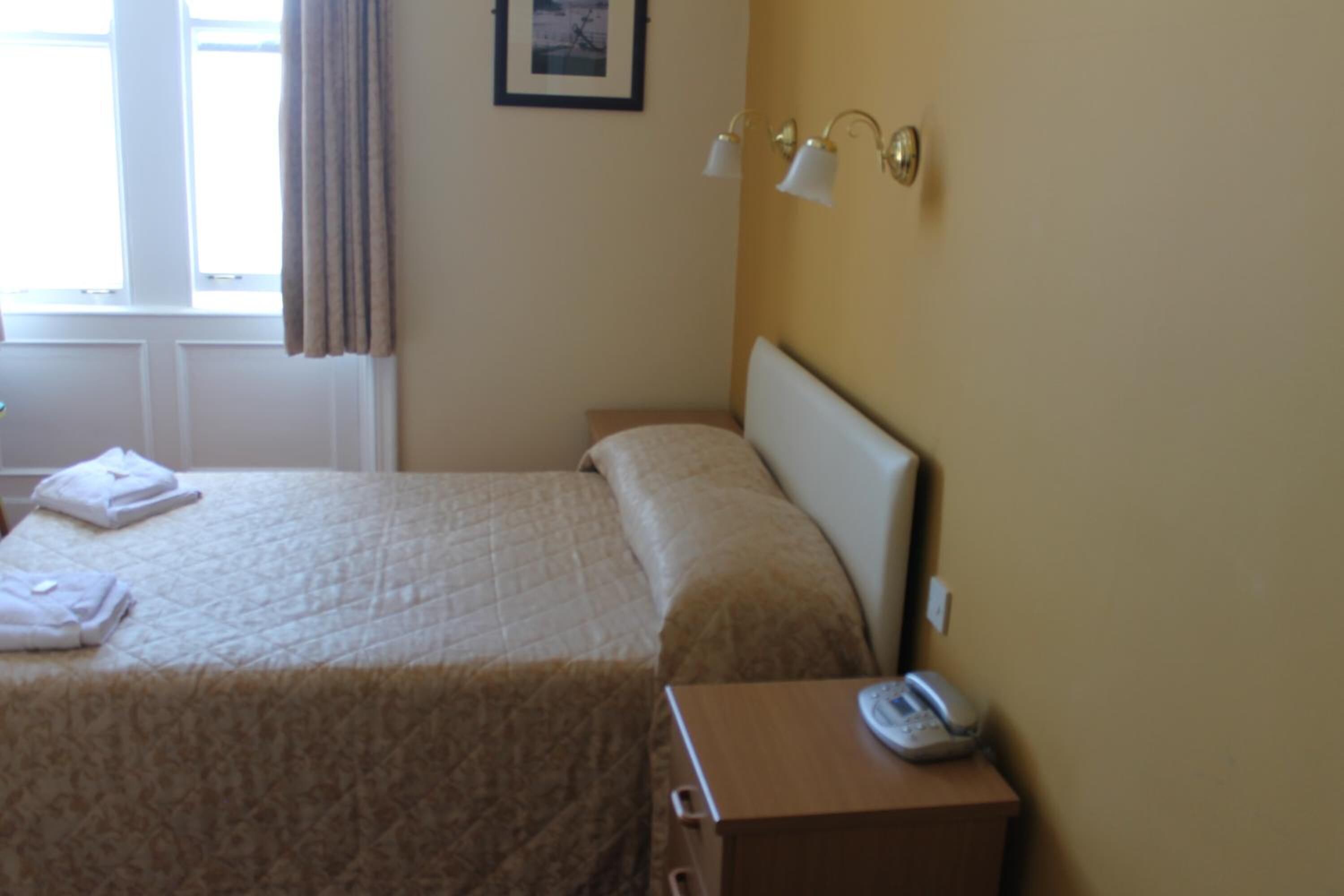 A hotel room with a bed, a bedside table with a telephone, and a window with curtains.