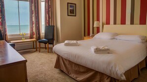 A hotel room with a bed, a desk, a chair, a window with a view of the sea, and a striped wall. - Chatsworth House Hotel (Llandudno)