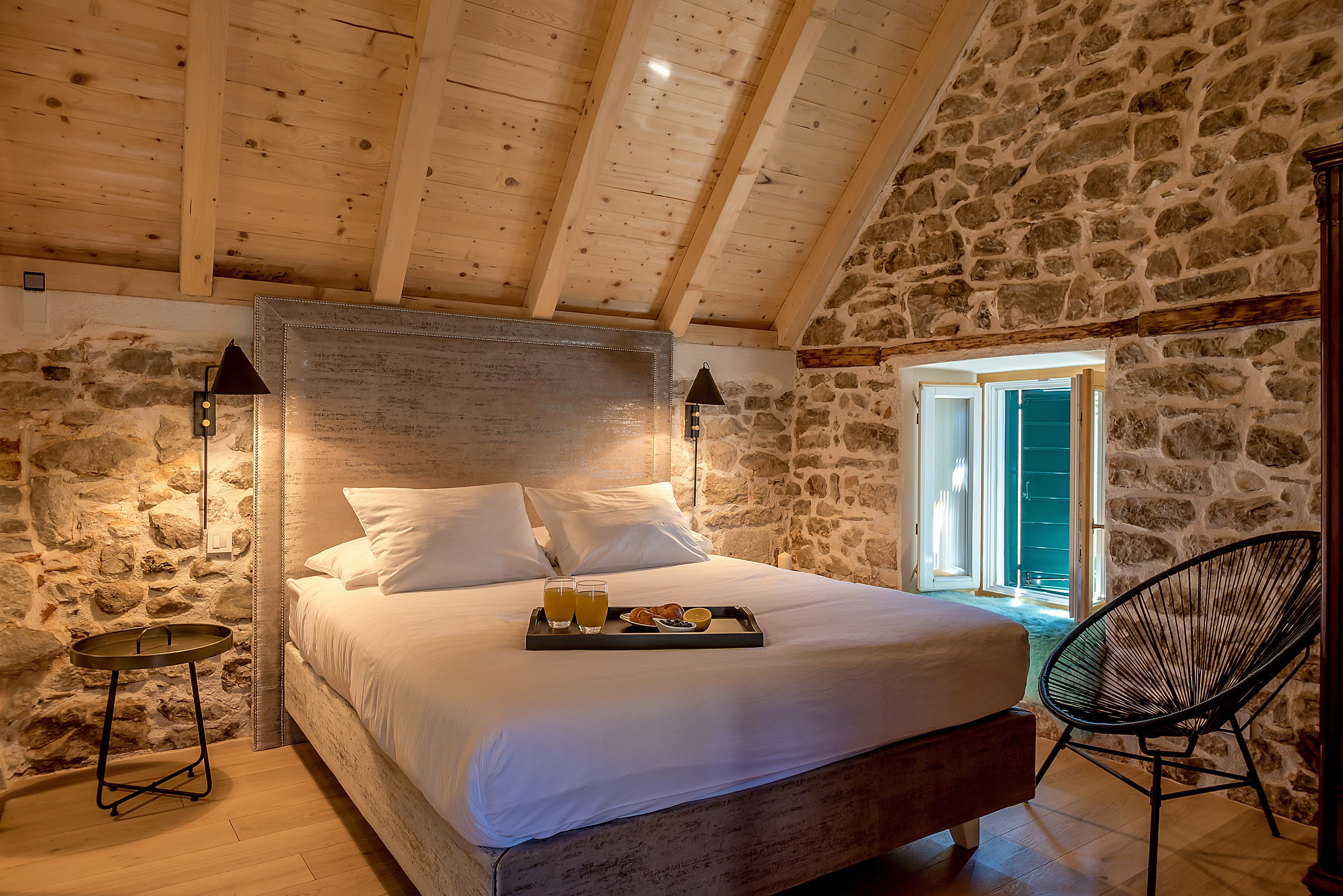 Une chambre avec un mur en pierre, des poutres en bois au plafond, un lit avec du linge de maison blanc, un plateau avec le petit-déjeuner et une chaise.