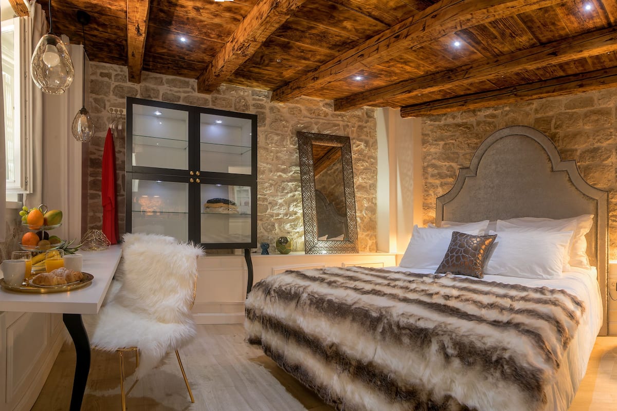 Un dormitorio con pared de piedra, techo de madera, cama con manta de piel, bandeja de desayuno y un espejo.