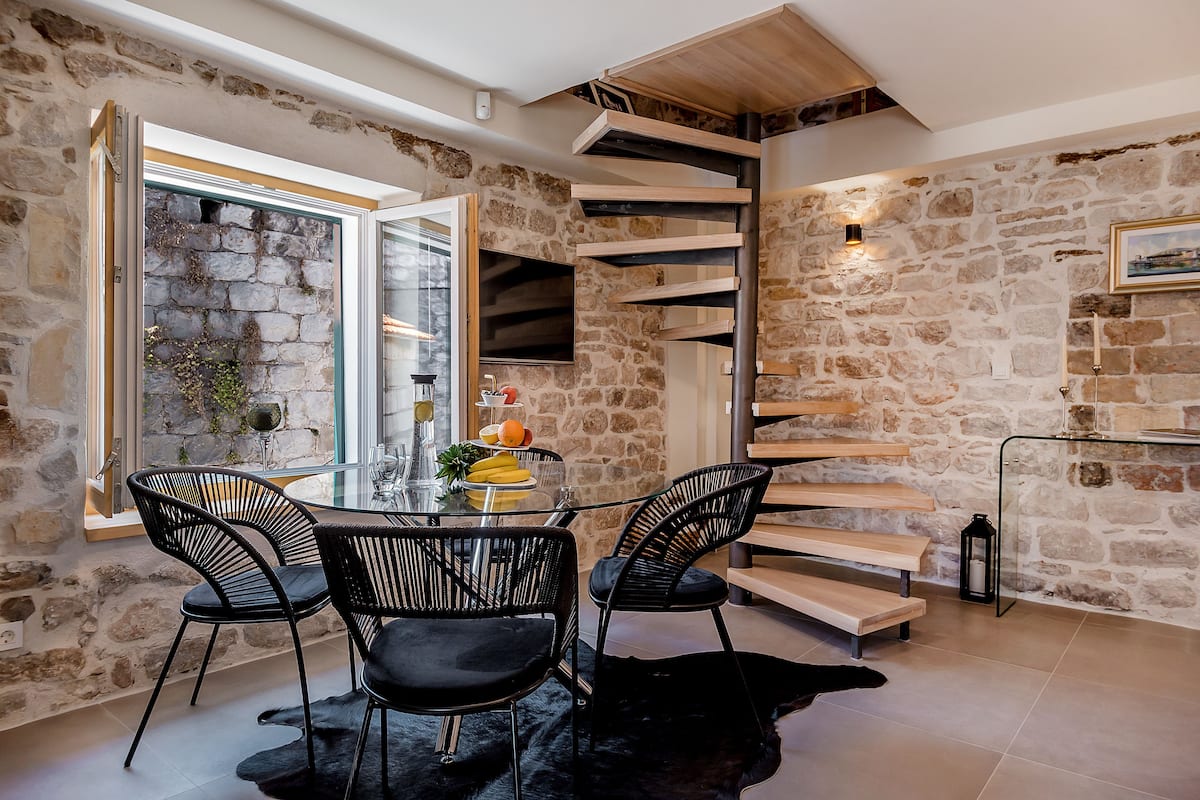 Un comedor con pared de piedra, escalera en espiral y mesa de cristal con sillas.