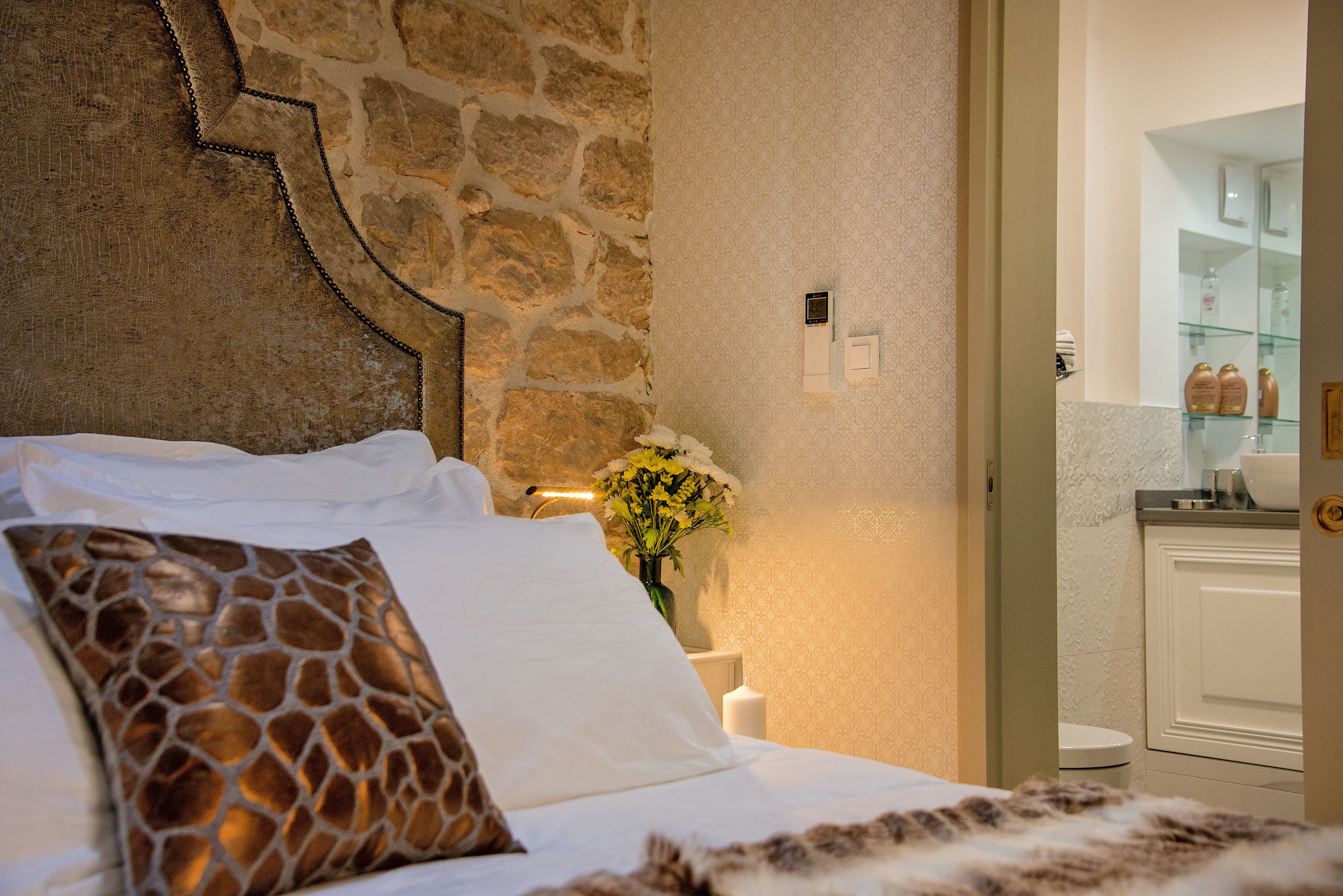 Una camera da letto con parete in pietra, un letto con biancheria bianca, un cuscino a motivi di giraffa e un vaso con fiori.