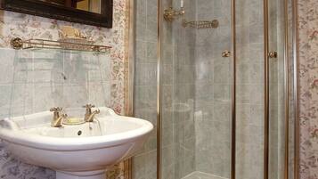 Un baño con un lavamanos blanco pedestal, una ducha con mampara de vidrio y papel tapiz con motivos florales.