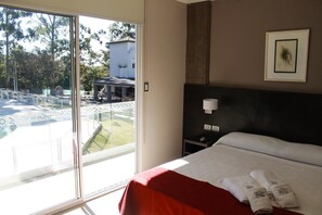 Ein Hotelzimmer mit Bett, Nachttisch, Wandlampe und Blick auf Pool und Außenbereich.