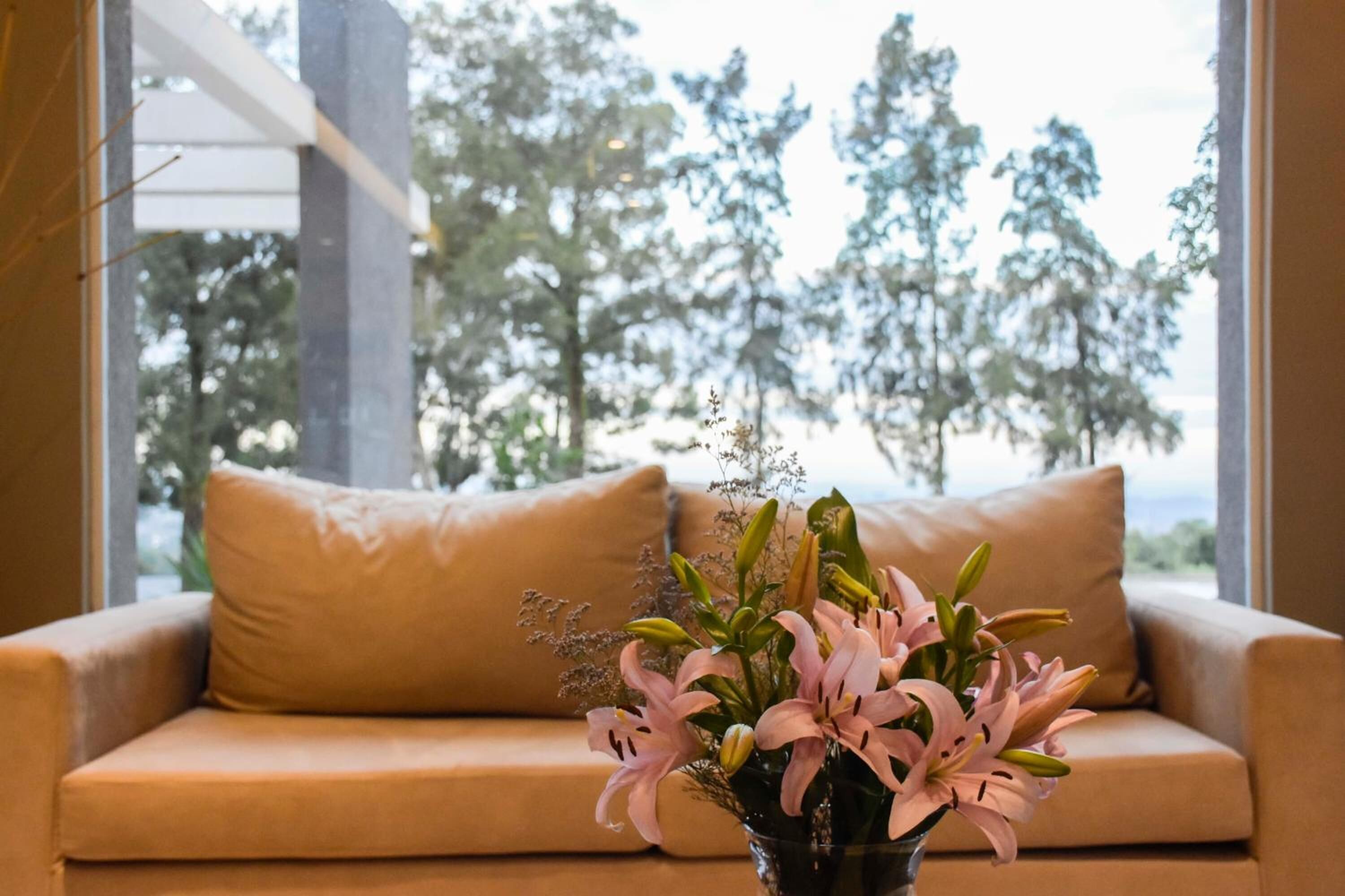 Um sofá bege com um vaso de lírios cor-de-rosa em frente a uma janela com vista para árvores.