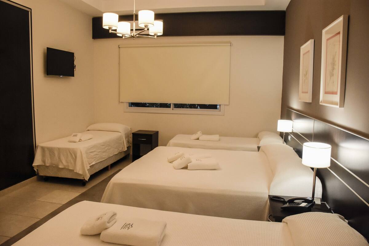 Habitación de hotel con dos camas individuales, una cama doble, televisión, mesita de noche con lámpara y ventana con persianas.