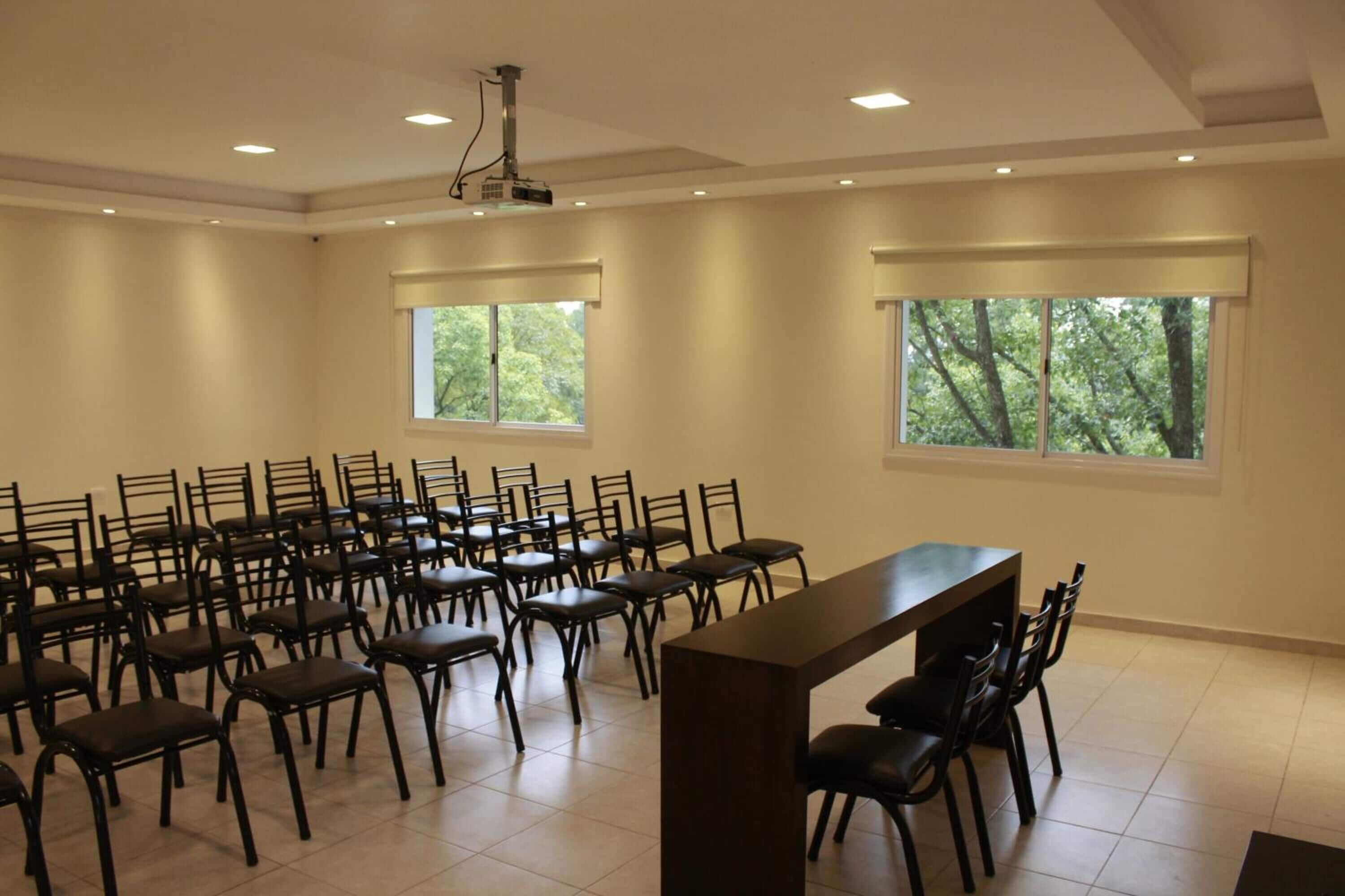 Un salón de conferencias con filas de sillas, una larga mesa y ventanas grandes con vistas a árboles.