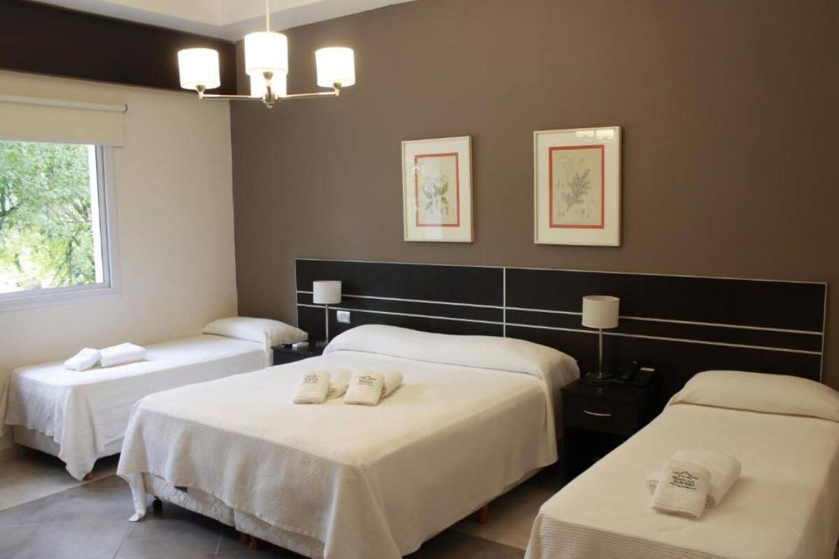Habitación de hotel con dos camas individuales, una cama grande y otra cama separada. Hay dos mesitas de noche con lámparas, una ventana con vista a árboles y dos cuadros en la pared.