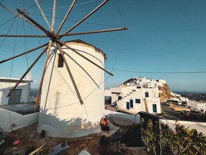 Un moulin à vent traditionnel, avec une base en pierre, entouré de bâtiments blancs aux portes et fenêtres bleues, sous un ciel bleu dégagé.