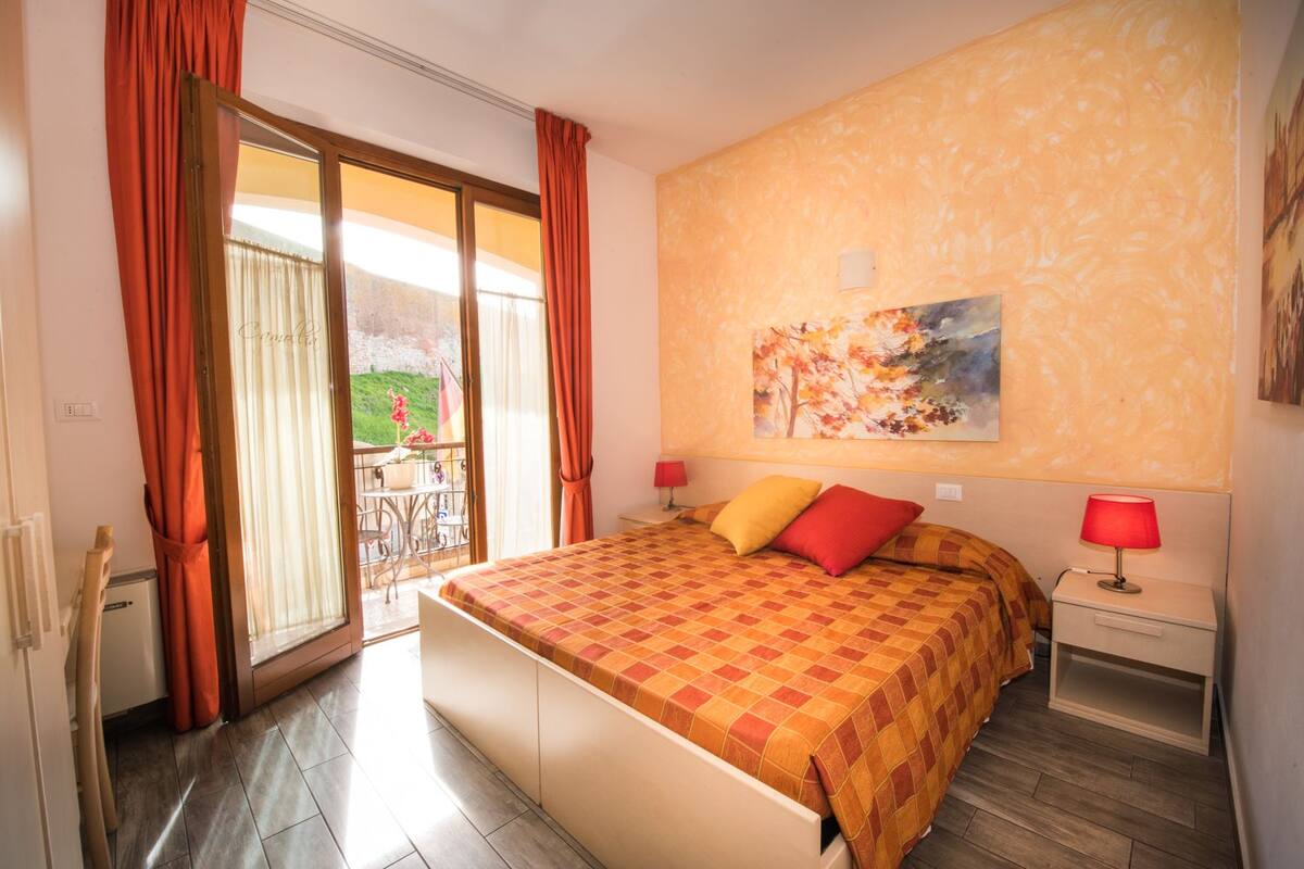 A hotel room with a bed, a nightstand, a lamp, a painting on the wall, and a balcony with curtains.