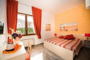 A bedroom with a bed, a desk with a chair, a window with curtains, a painting on the wall, and a lamp on a bedside table. - B&B Camollia (Siena)
