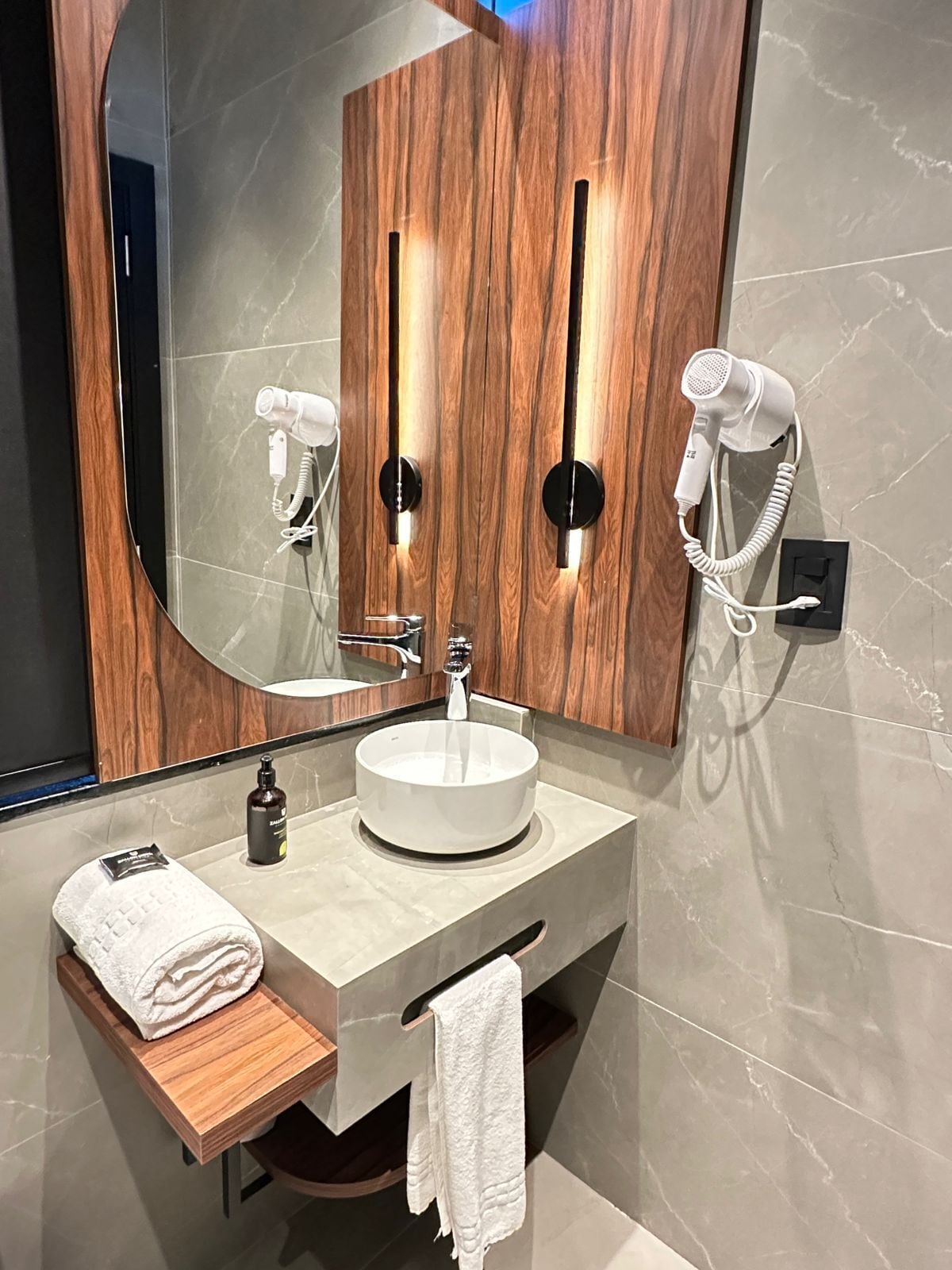 A modern bathroom with a round sink, wooden vanity, a large mirror, and two hair dryers mounted on the wall.
