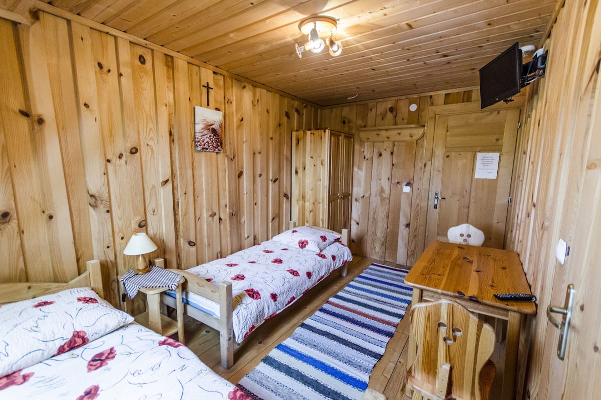 Una habitación tipo cabaña de madera con dos camas individuales, una mesita, una lámpara y un televisor fijado a la pared.