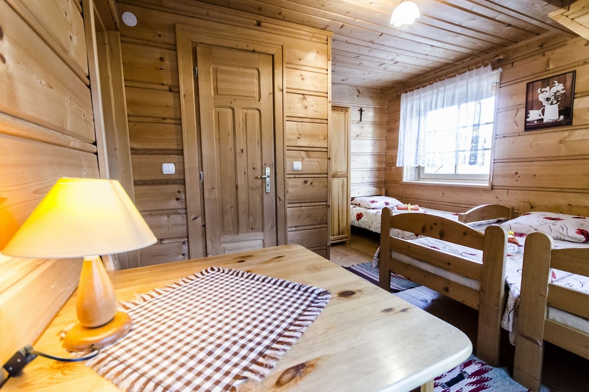 Una habitación de cabaña de madera con una cama, una mesa con lámpara y una ventana con cortina.