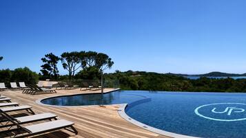 Une magnifique piscine à débordement d’un bleu profond, ornée du logo UP, entourée d’une terrasse en bois et de chaises longues, et offrant une vue imprenable sur un paysage pittoresque avec des collines et un lac.