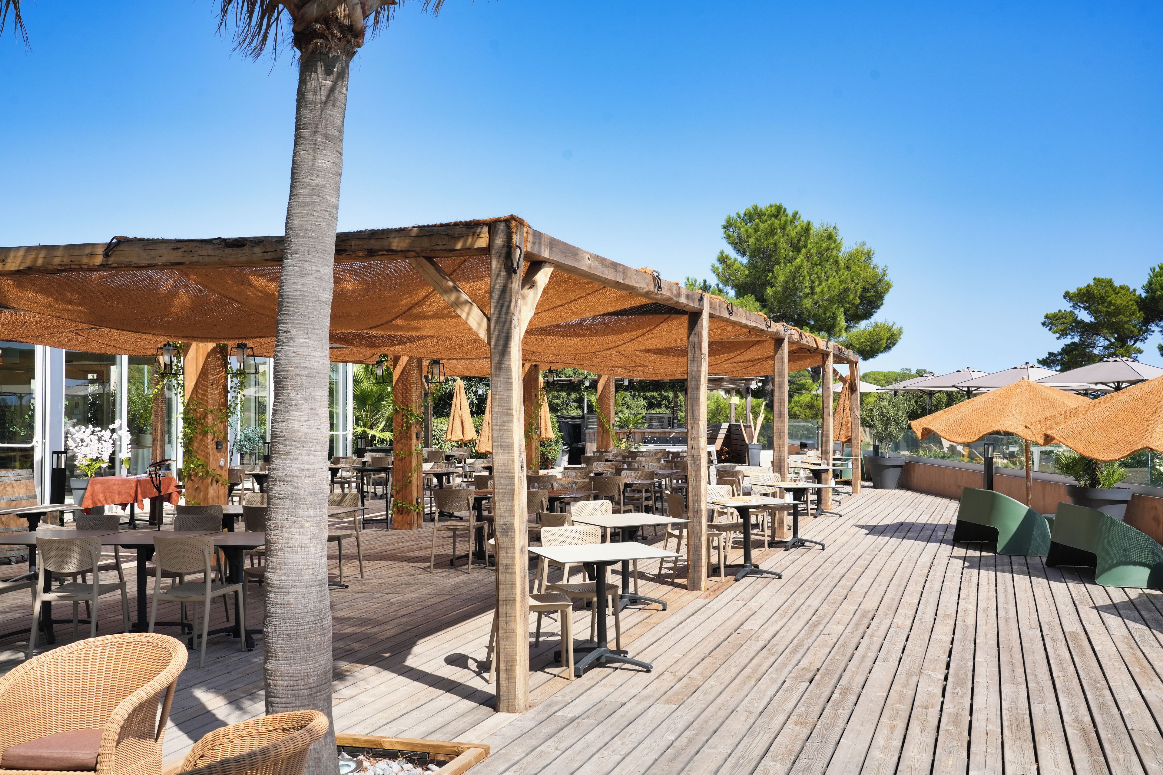 Un área de patio al aire libre con mesas y sillas de madera, cubierta por una pérgola con sombrilla tejida.