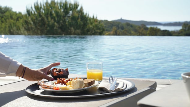 A breakfast tray with eggs, bacon, toast, and orange juice on a table by a lake. - Hôtel U Paesolu (Zonza)