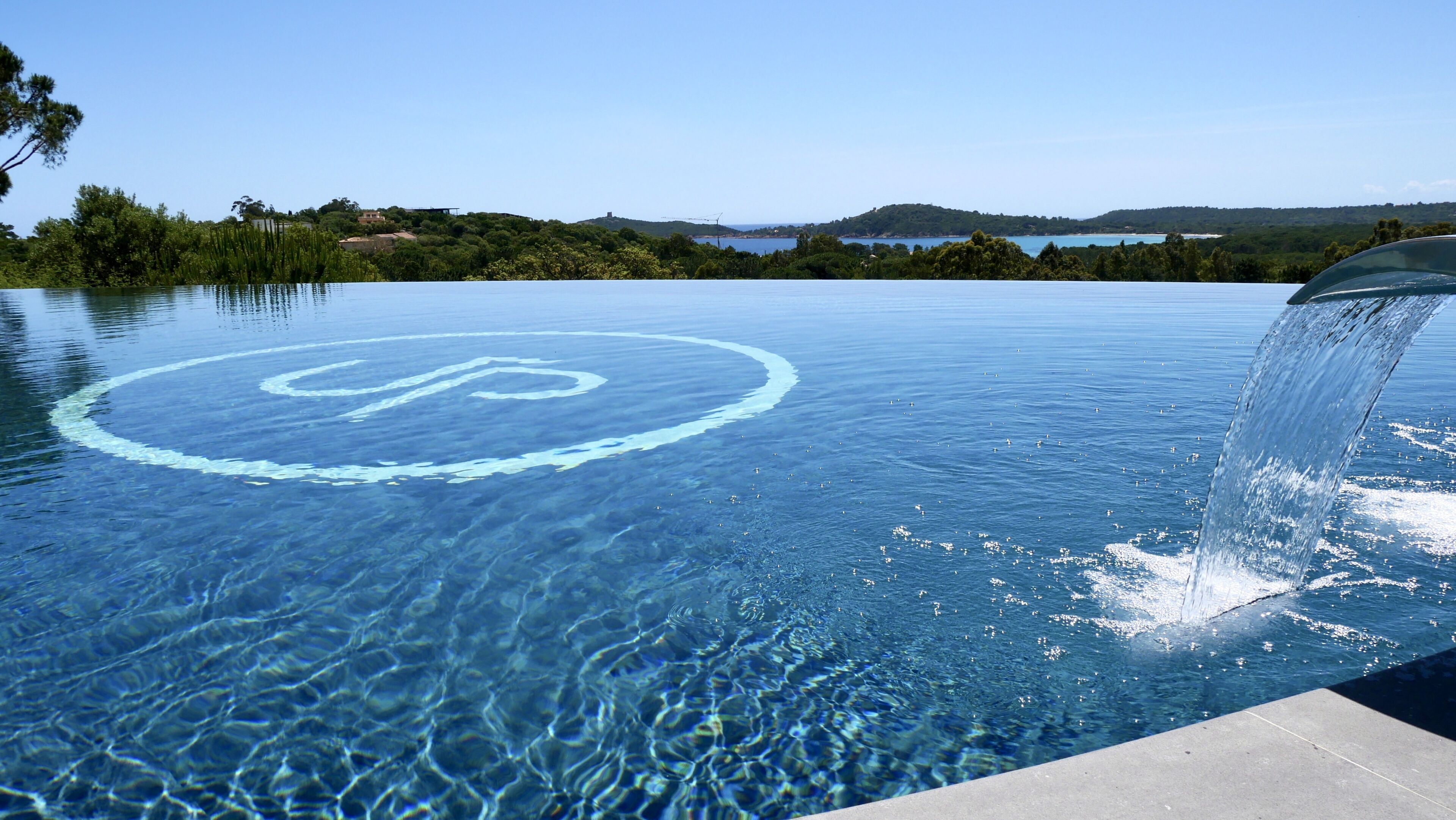 A clear blue infinity pool with a circular logo, surrounded by a natural landscape and a view of hills and water.