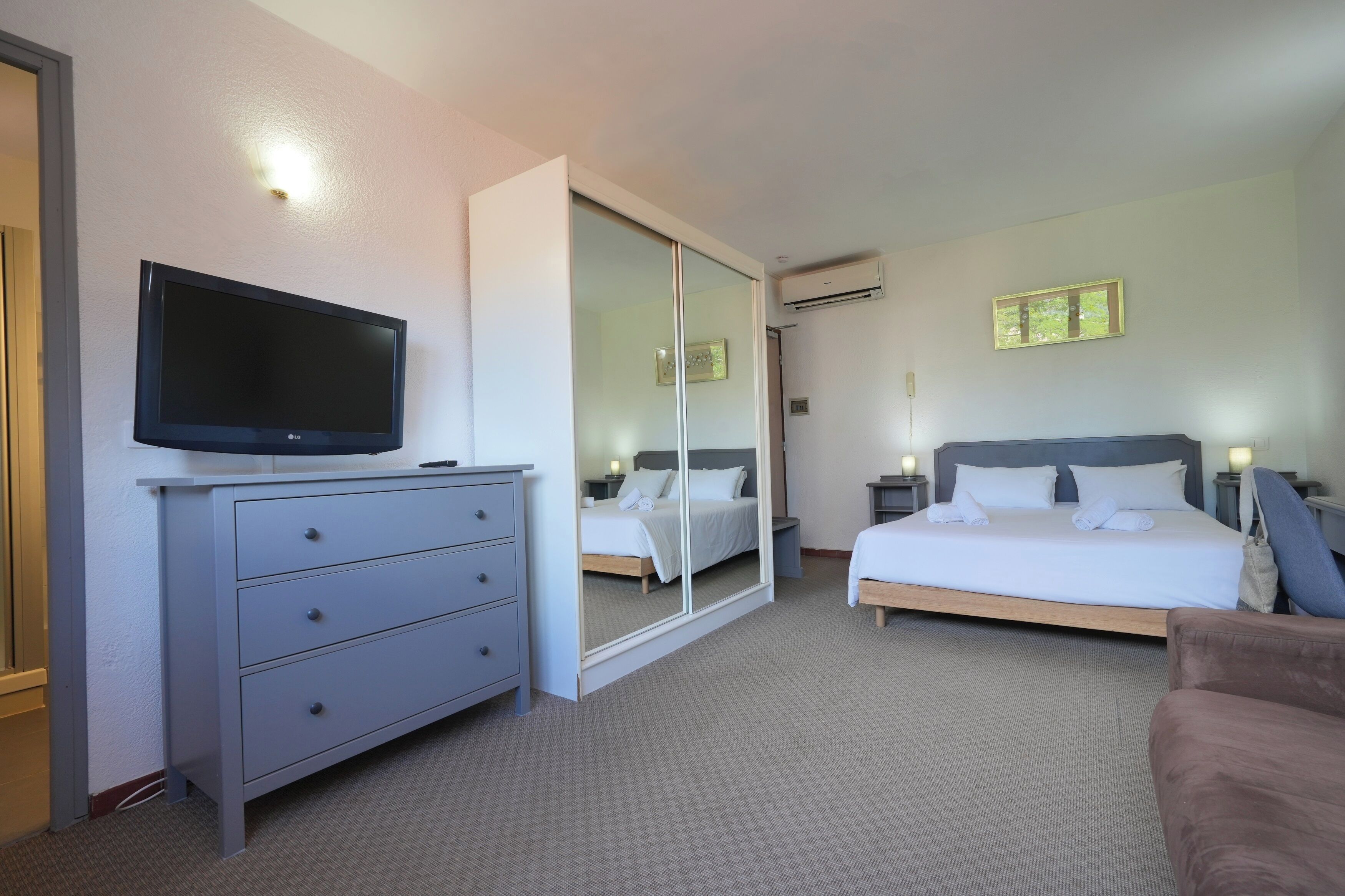 A hotel room with a flat-screen TV on a dresser, a large mirror, a bed with white linens, a bedside table with a lamp, and an air conditioning unit on the wall.