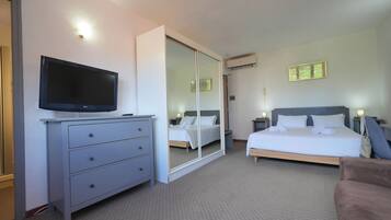 A hotel room with a flat-screen TV on a dresser, a large mirror, a bed with white linens, a bedside table with a lamp, and an air conditioning unit on the wall.