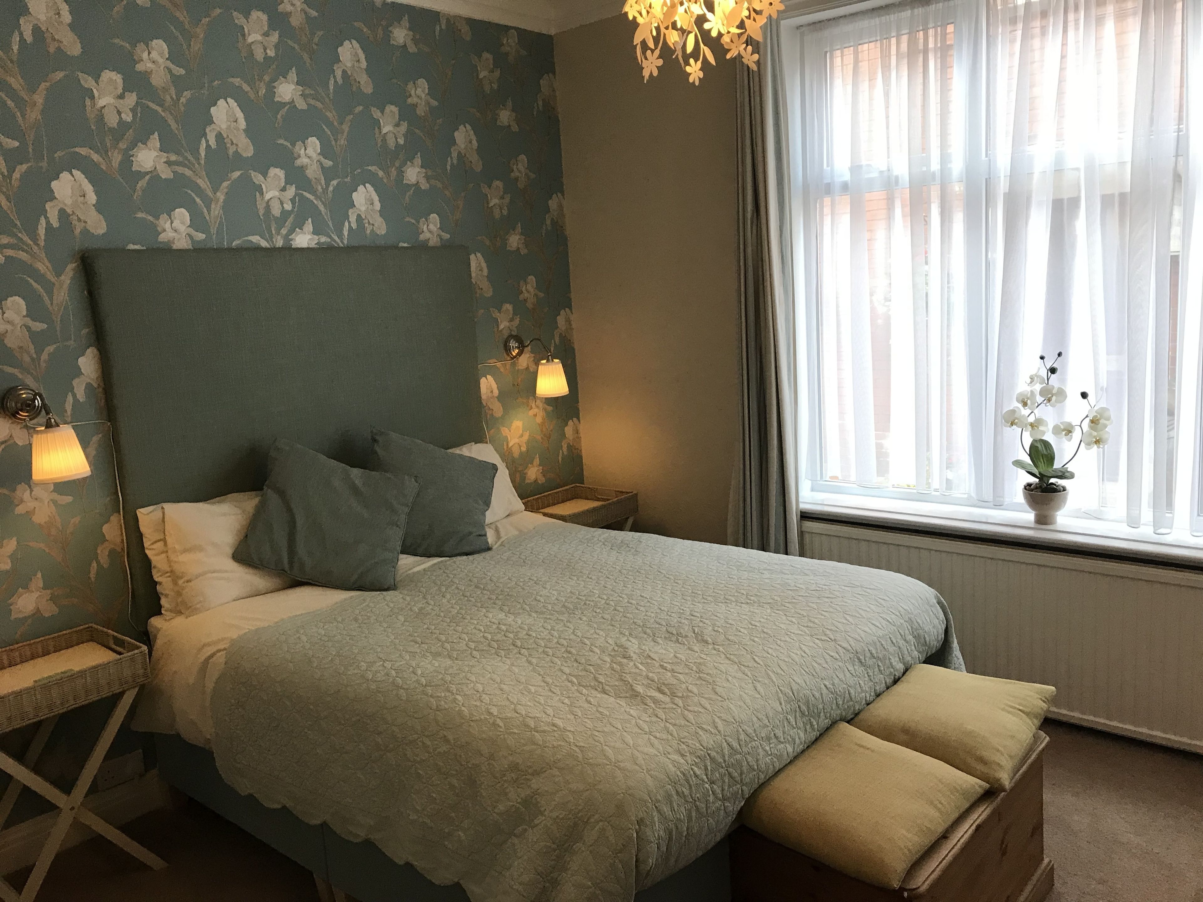 A bedroom with a large bed, two bedside tables, a nightstand, a window with sheer curtains, and a small potted orchid on the windowsill.