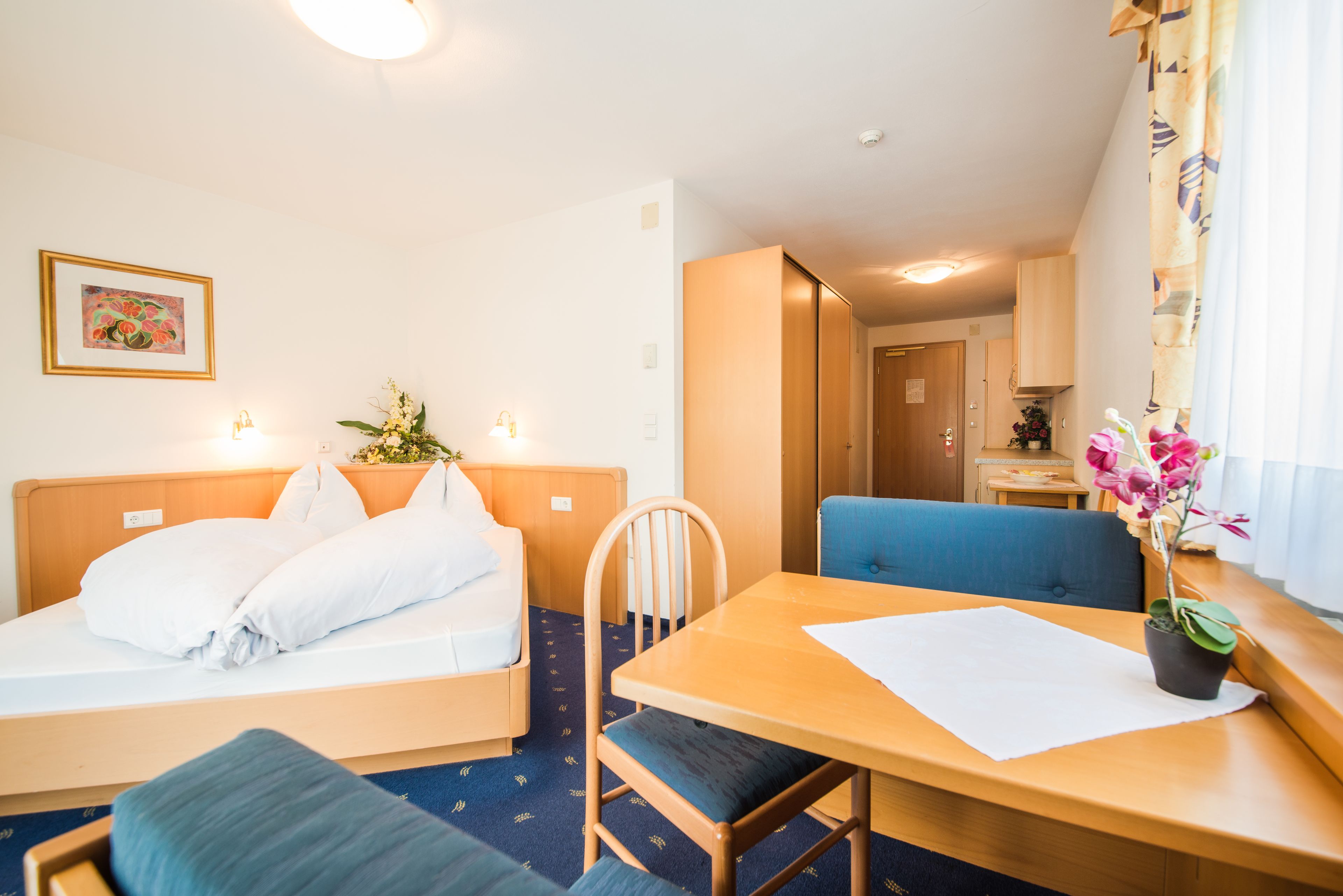 A hotel room with a bed, a table, a chair, a small table with a plant, and a window with curtains.