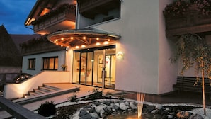Entrada de hotel con puerta de cristal, iluminación exterior y una pequeña fuente de agua.