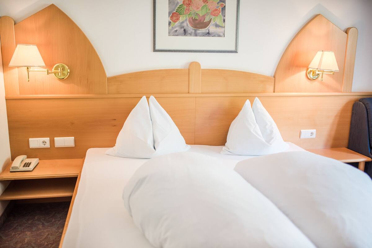 A hotel room with a wooden headboard, two bedside lamps, a telephone, and a framed floral artwork on the wall.