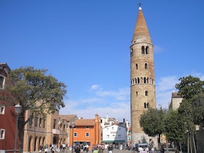 A historic tower with a conical roof and arched windows, surrounded by traditional buildings and a busy square. - Hotel Catto Suisse (Caorle)