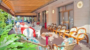 An outdoor seating area with wicker chairs and tables, a covered walkway, and a building entrance. - Hotel Catto Suisse (Caorle)