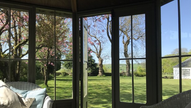A conservatory with a view of a well-maintained lawn and trees, a wicker sofa, and a door. - South Craighall Bed & Breakfast (Glasgow)