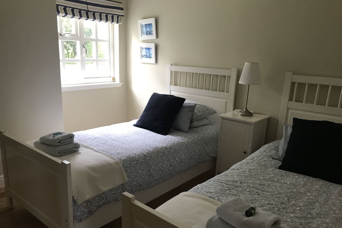 A twin room with two beds, a window with curtains, a bedside table with a lamp, and a wall with framed pictures.