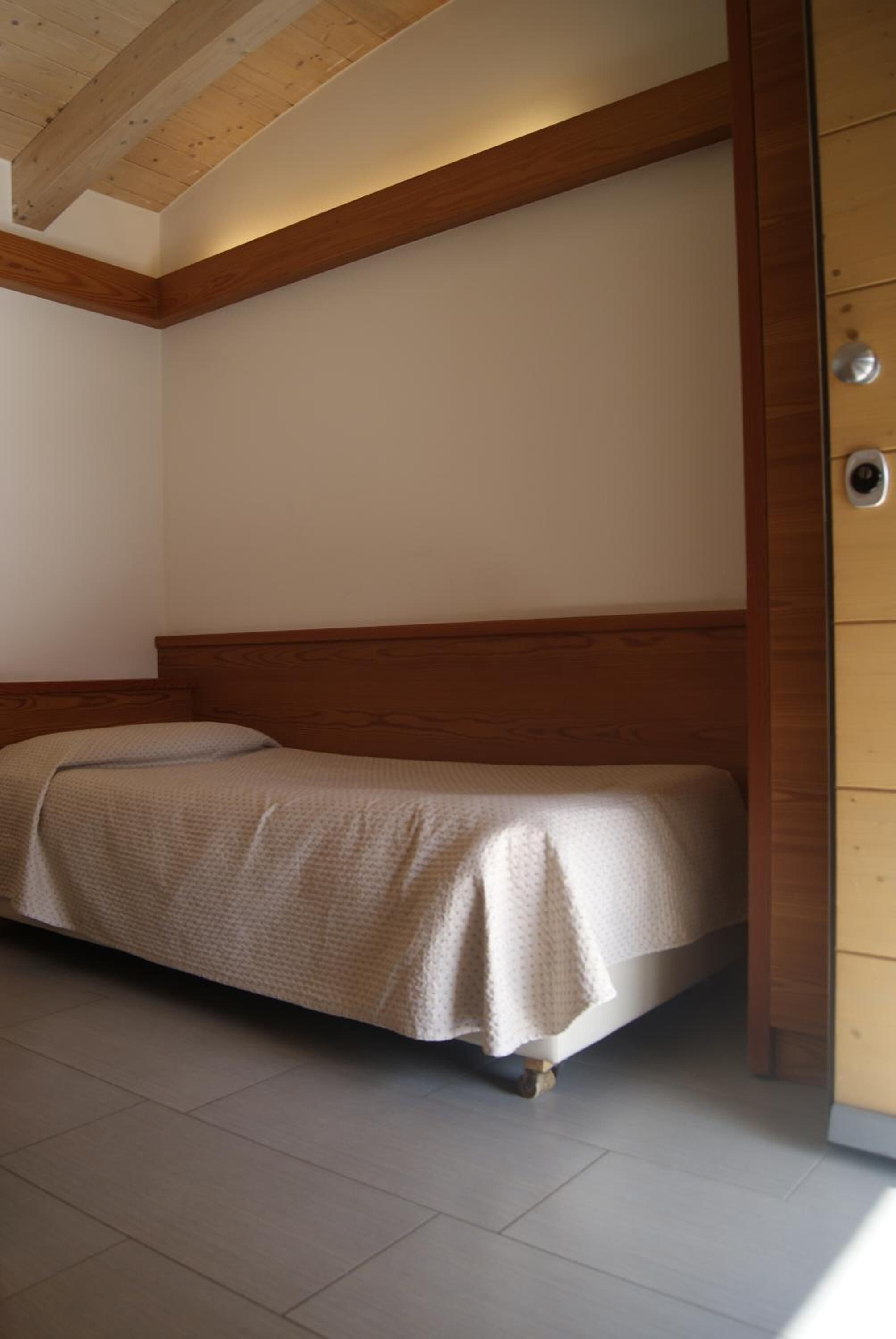 Un lit simple avec une tête de lit en bois, dans une pièce aux murs blancs et dotée d’une poutre en bois au plafond.