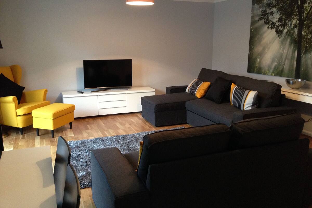 A modern living room with a grey sofa, yellow armchair, and ottoman, a flat-screen TV on a white cabinet, and a large abstract painting on the wall.