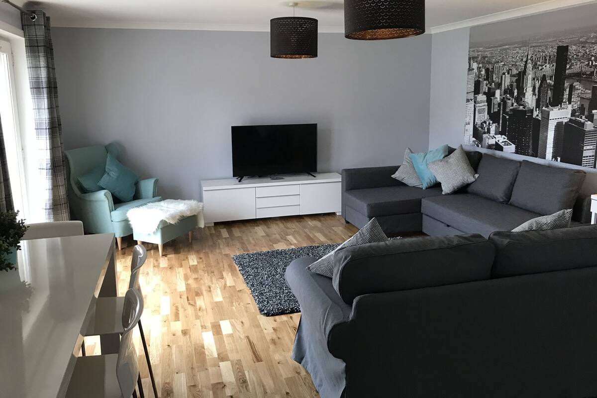 A modern living room with a grey sofa, a teal armchair, a flat-screen TV on a white console, and a cityscape wall mural.