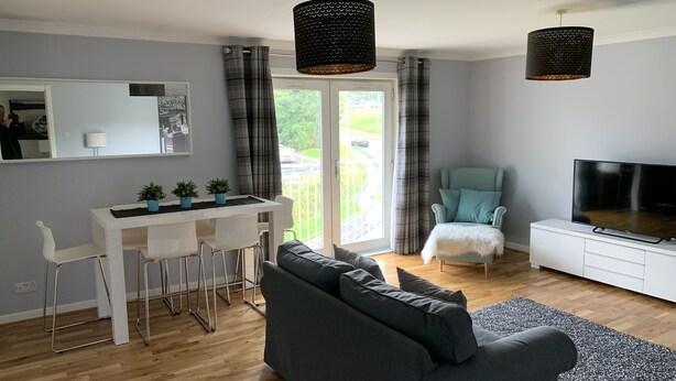 A modern living room with a grey sofa, a dining area with white chairs, a TV on a white stand, and a balcony with curtains. - Serviced Apartments East Kilbride (Glasgow)